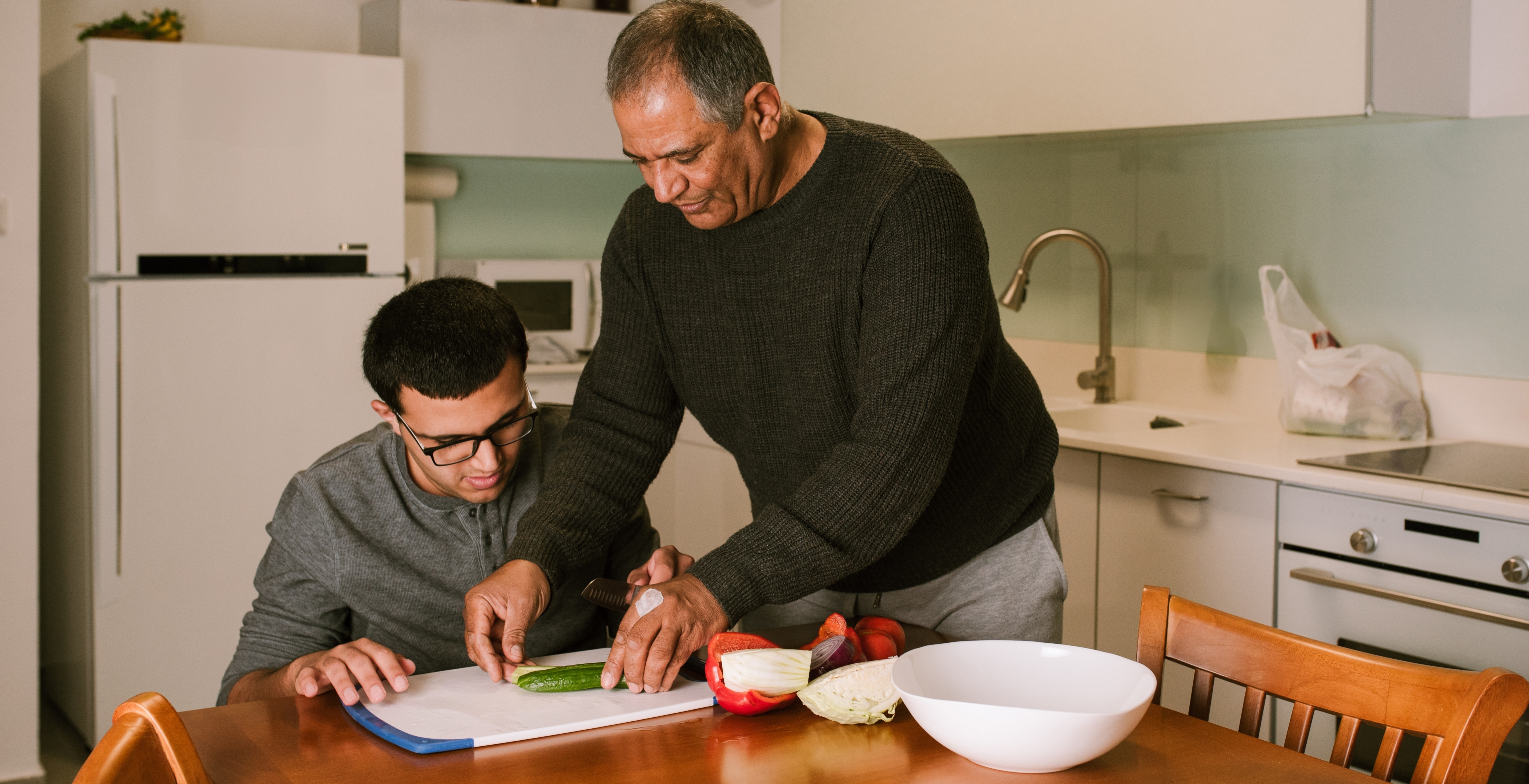 Older man helping teenage song cut food on a cutting board