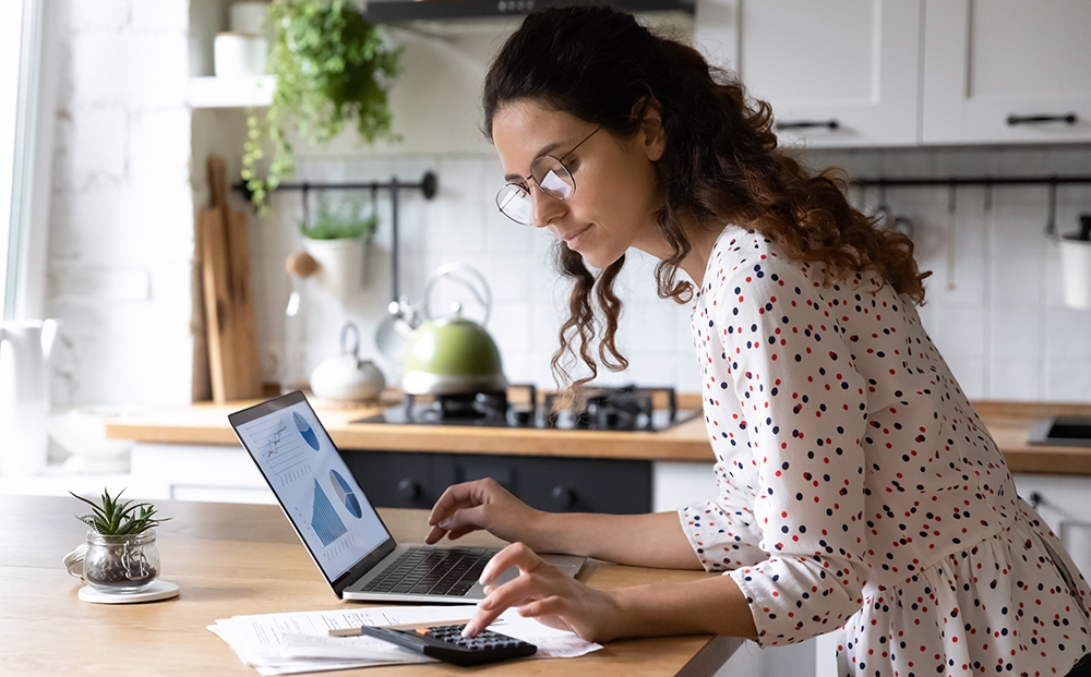 woman sitting at kitchen table looking at computer and paperwork while using a calculator