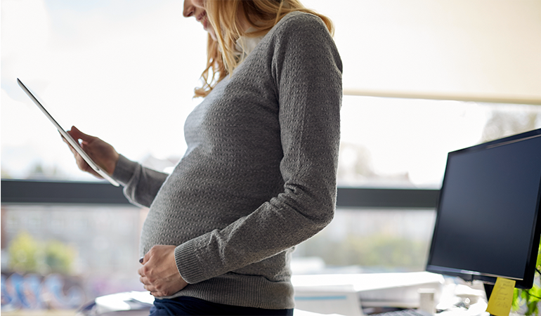Pregnant businesswoman stands in office while looking down at tablet with one hand resting on her belly.