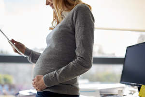 Pregnant businesswoman stands in office while looking down at tablet with one hand resting on her belly.