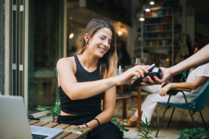 Young woman sitting in a cafe in downtown district paying by card for a coffee.