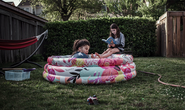 Young girl in a kiddie pool with another older girl reading behind her.