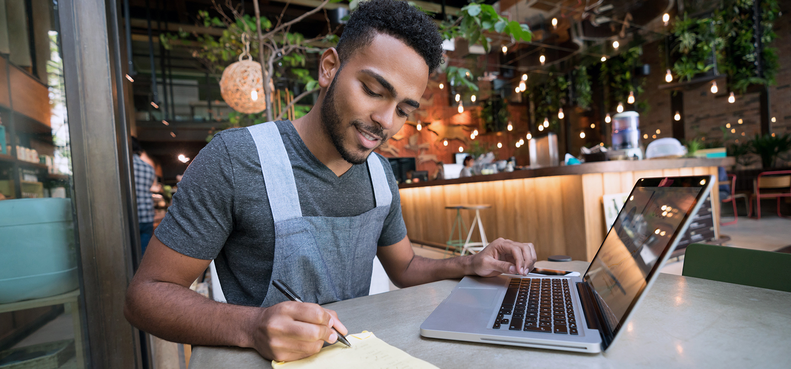 Young male business owner wearing an apron in a cafe writing down notes while working on a computer