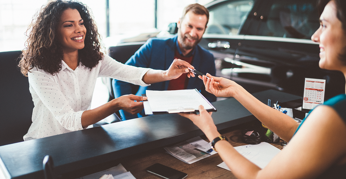 Woman handing over a clipboard of documents to a woman at a dealership