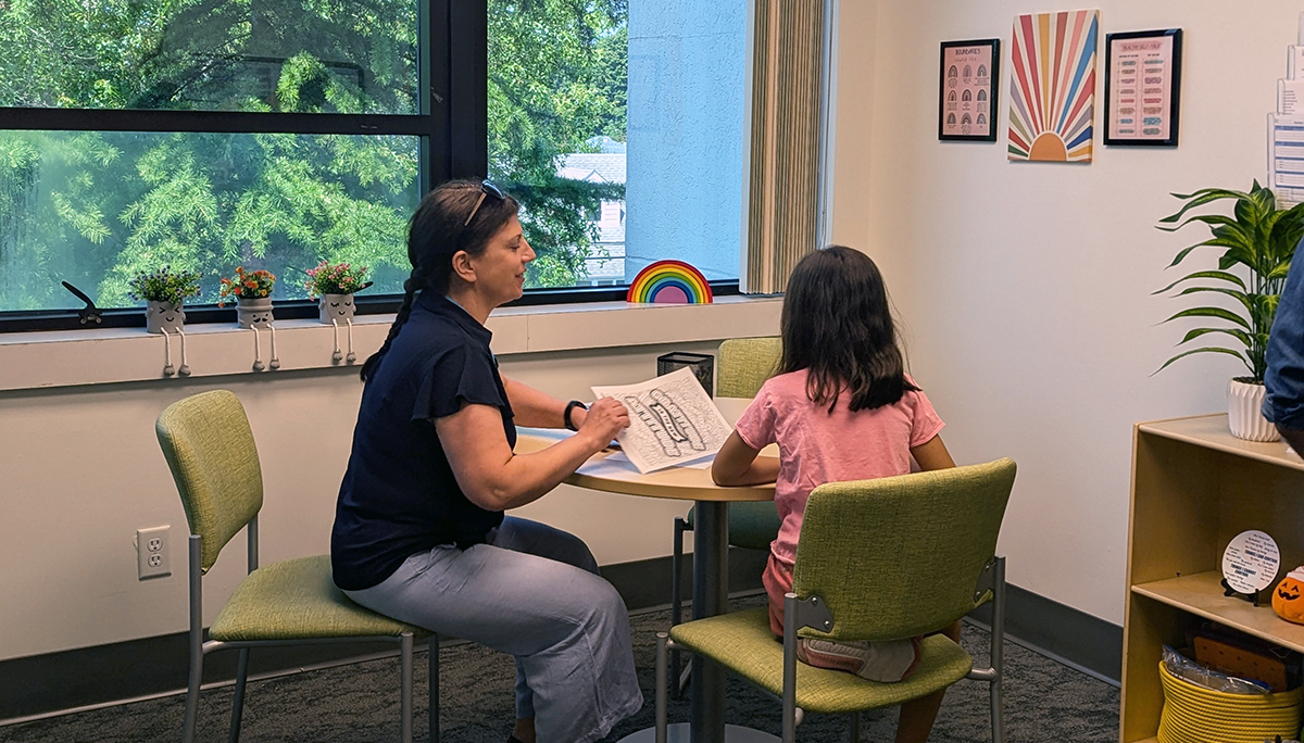 Woman teacher sitting at a desk with a young girl, coloring pictures