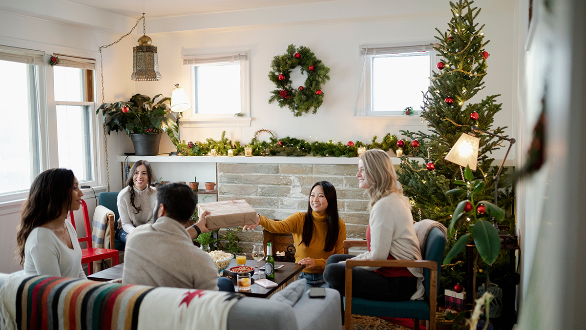 Group of adults sitting around a living room decorating with holiday decorations passing around holiday gifts