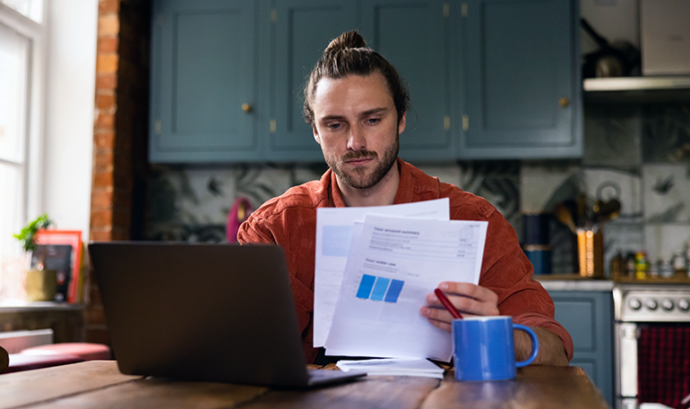 Young adult man holding documents while looking at a laptop