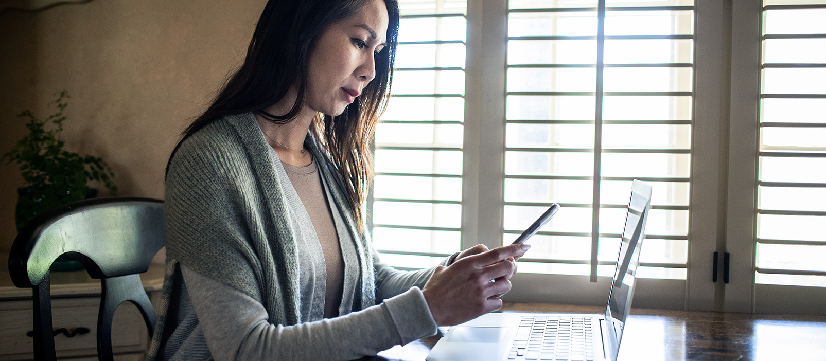 Woman looked stressed sitting at a kitchen table with laptop open in front of her, and looking at a cell phone