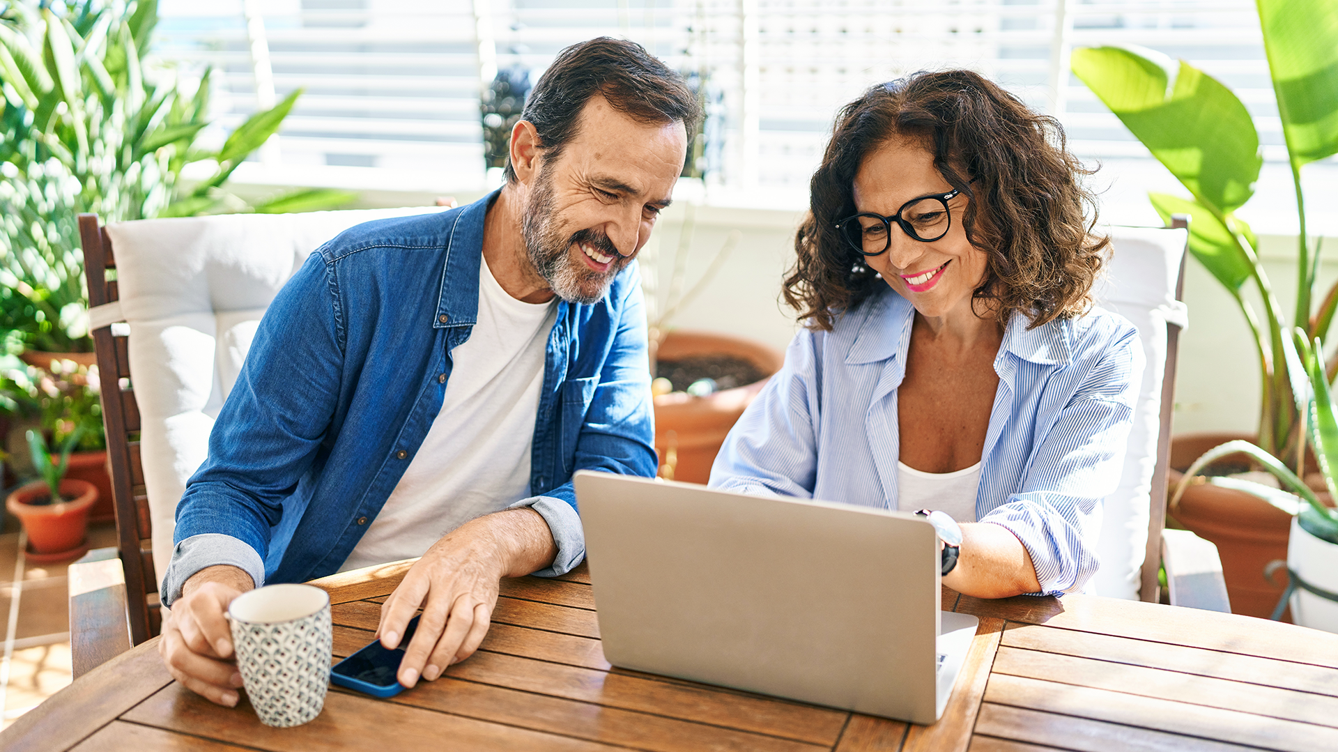Older woman on laptop sitting at a kitchen with man sitting next to her, looking at the laptop, holds a cup of coffee