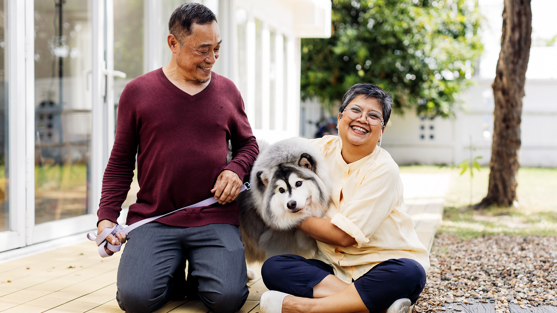 Older woman sitting on the ground hugging a white and gray fluffy dog while man holds the leash smiling