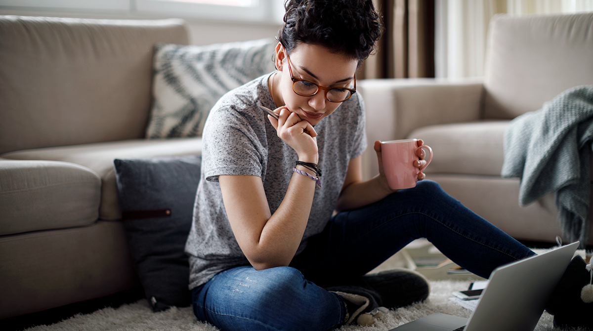 Woman sitting on the floor with a cup of coffee looking deep in thought while looking at a laptop