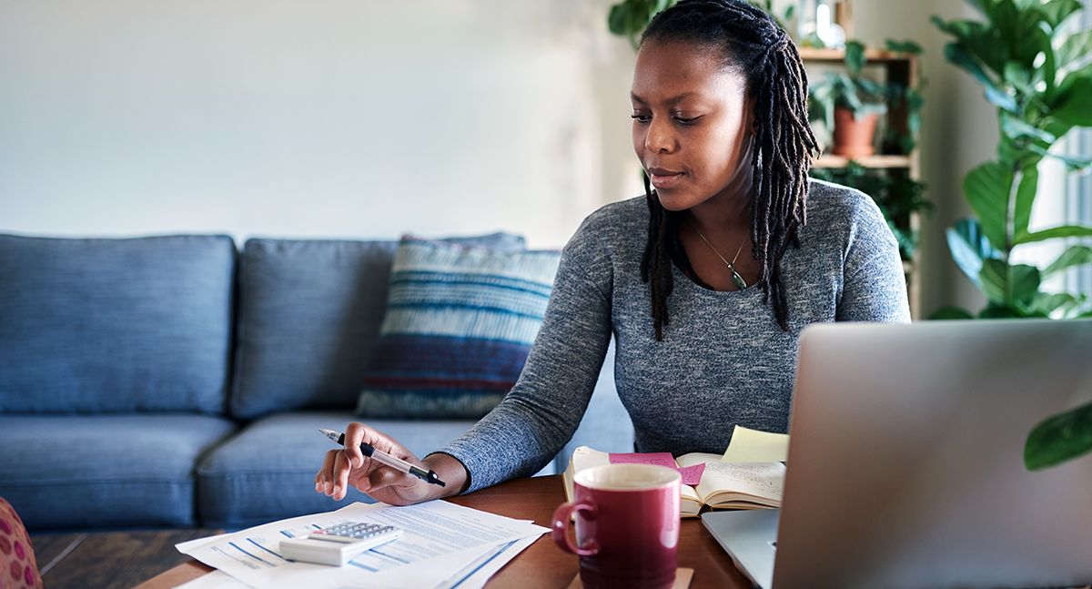 Woman at home looking at paperwork and using a calculator to do her taxes on a computer