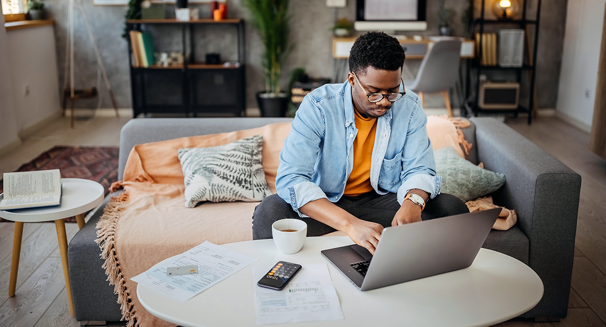 Young man at home sitting on a couch working on a laptop on table that had documents on it