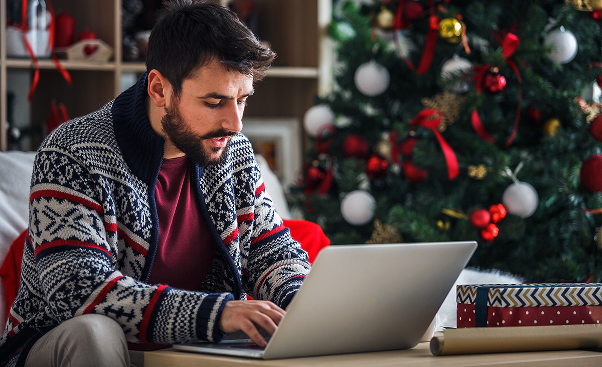 man at home with holiday decor around him wearing a holiday sweater on a laptop