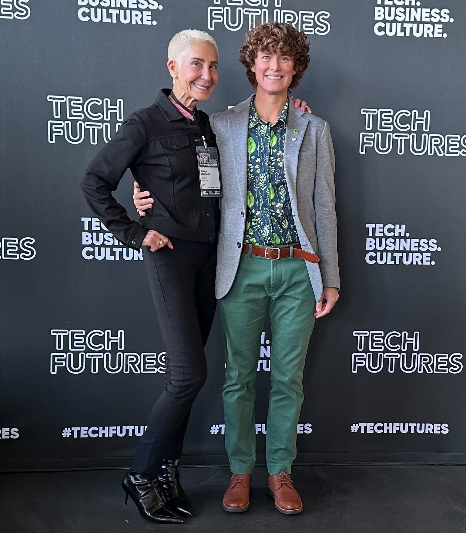 Robyn Streisand (left, woman with short white hair and black top and pants) with Jennie Platt (woman with short, curly brown hair, with green pants, a patterned button down, and a gray blazer) in front of a Tech Futures banner