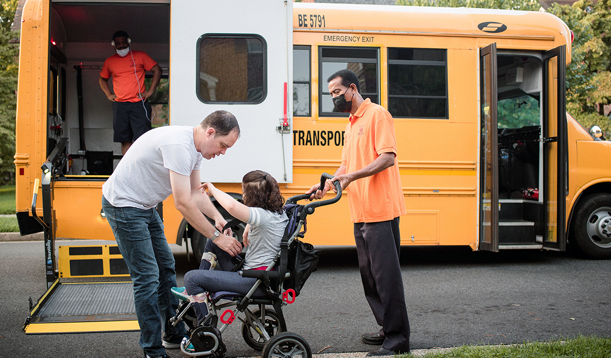 Young girl in wheel chair being helped by two men to get on a school bus