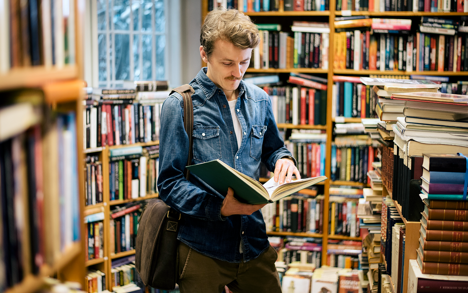 Man in a library, wearing a shoulder bag, reading a book